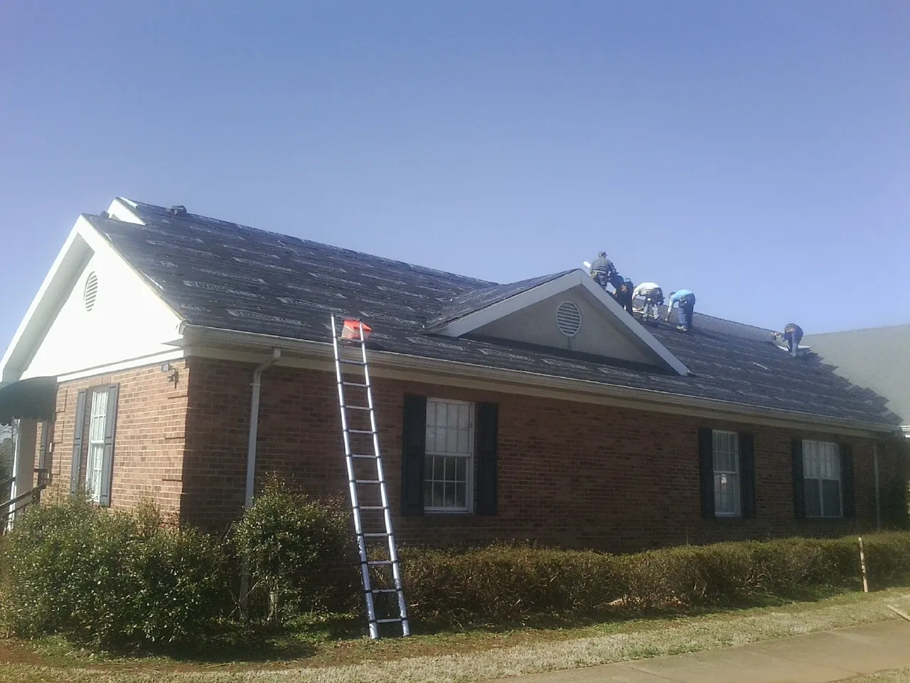 Crew tearing shingles off a brick ranch roof with a ladder set — Emergency Roof Repair work by our crew in New Concord