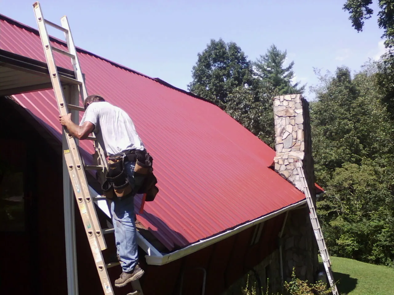 Roofer on a ladder beside a red metal roof and stone chimney — Soffit & Fascia Repair by Roofing Brothers in New Concord